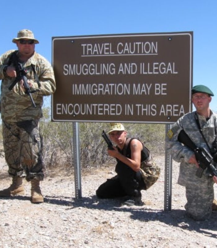 Image of three Arizona Minutemen posing with their rifles around a sign about illegal smuggling and immigration